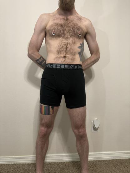 A person standing against a light-colored wall, wearing black Under Armour boxerjock. Their torso is exposed, displaying chest hair, two nipple piercings, and a tattoo on the left arm. A multiple pride flag tattoo is visible on their upper right thigh.