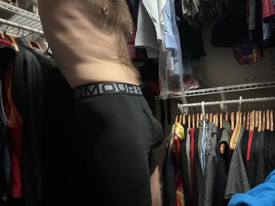 A person in black Under Armour standing in a walk-in closet filled with hanging clothes.