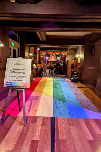 A rainbow carpet leading to the entrance of the Hearthstone Lounge at the Grand Californian Hotel.

A sign reads:
Disney's 
GRAND CALIFORNIAN
Hotel & Spa

Come join us for Disney Pride

HEARTHSTONE LOUNGE

June 18th & 20th 4:00pm to Midnight

Reservations are recommended as space is limited.
Walk-ins are welcome!