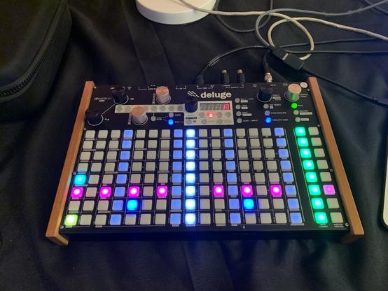 A music production device with multiple buttons and knobs, displaying colorful LED lights. The word "deluge" is prominent on the device.