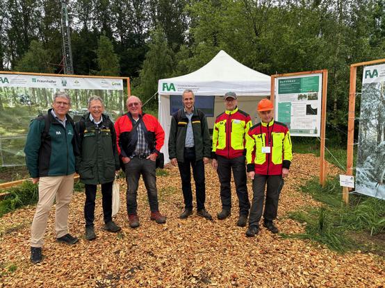 Forstfachleute auf einem Informationsstand im Wald. Im Hintergrund informieren Poster und Banner über wissenschaftliche Projekte.