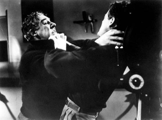 In this image, there are two men in a tense confrontation. The man on the left is grabbing the collar of the shirt worn by the man on the right, and his facial expression suggests he might be angry or aggressive. The man on the right appears to be struggling against this forceful grip. They are both dressed in dark clothing, which adds a dramatic effect to the scene. There's no text visible in the image, and the background is blurred, making it difficult to discern specific details about their surroundings. The overall tone of the image suggests an intense moment from a film or drama.