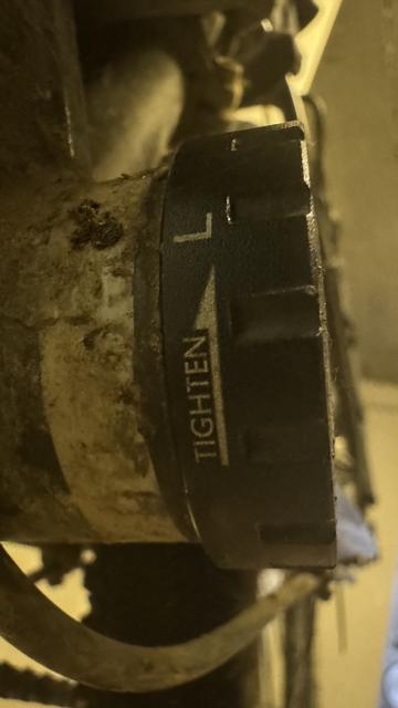 Nahaufnahme eines Metallrohrs mit einer schwarzen Kappe mit der Aufschrift "TIGHTEN" und einem Pfeil, der die zu drehende Richtung anzeigt. Das Bild zeigt eine abgenutzte und schmutzige Oberfläche.