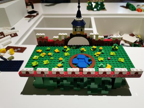 Ein Modell aus Klemmbausteinen auf einem weißen Tisch.
Es ist ein Garten mit Teich umrahmt von kleinen Bögen. der hintere Teil besteht aus einem großen Bogen mit Turm.