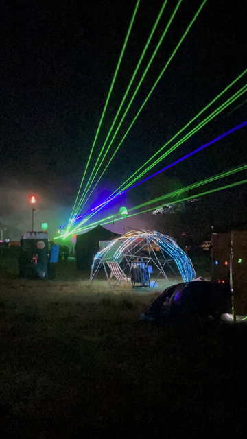 Photo showing a campsite after dark. Multiple green and blue laser beams are emanating from a central point. In the foreground is an illuminated geodesic dome with several deckchairs and a generative music console. To the left hand side is a Datenklo "data toilet". Pumping techno and flamethrowers are left to the imagination...