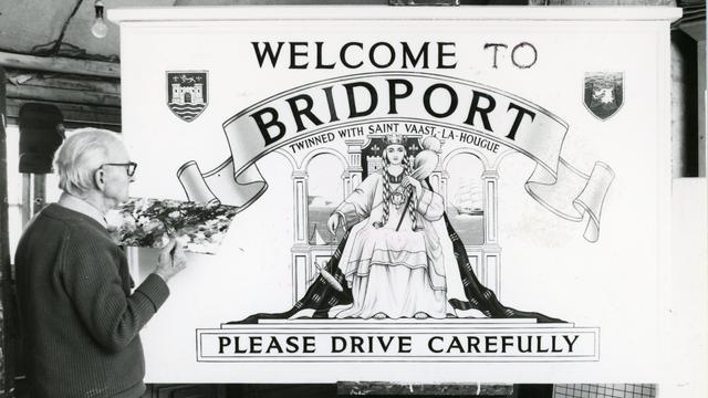 Black and white photo of an elderly man with a painter's palette standing in front of a 'Welcome to Bridport' sign that he's working on.

The sign features a combination of lettering and pictorial elements, which includes the text 'Twinned with Sant Vaast-la-Hougue' and 'Please Drive Carefully'.

The picture is of a queen on a throne in a flowing dress and gown, and arched windows behind with sea views.

There are also two coats of arms flanking the main lettering at the top.
