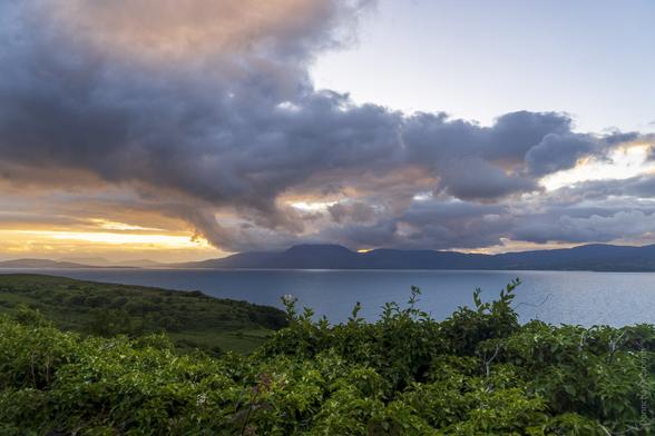 A distant sunset with clouds and the sun lighting up the sky. Distant hills are separated from the viewer by water and in the foreground are bushes of green leaves.