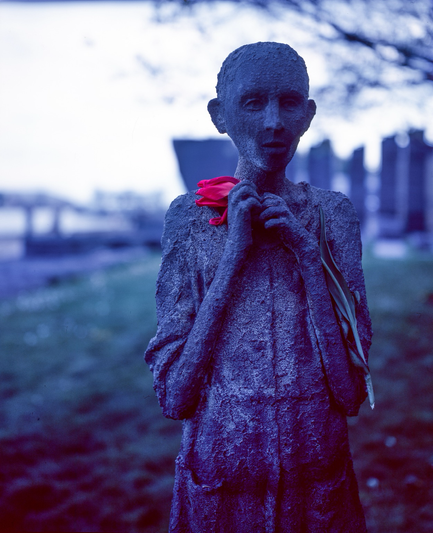 A stature in memory of the refugees of the Irish Famine arriving in Toronto, a thin and gaunt child holds their hands together near the base of their neck, nervous to see a new land. Someone has added a red flower to the statue and it appears the child is clutching it. The old film used has added a blue tone to the image and the flower is brilliant red.