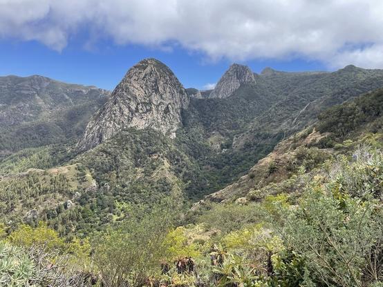 Blick auf den Roque Cano in Vallehermoso. Man sieht grün bewachsene Hänge, der Himmel ist blau mit größeren Wolkenbereichen