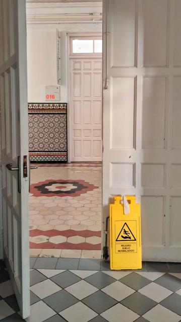 A través de una puerta entreabierta se ve un pasillo y otra puerta. Al lado de la puerta hay un cartel amarillo que pone "peligro suelo resbaladizo"