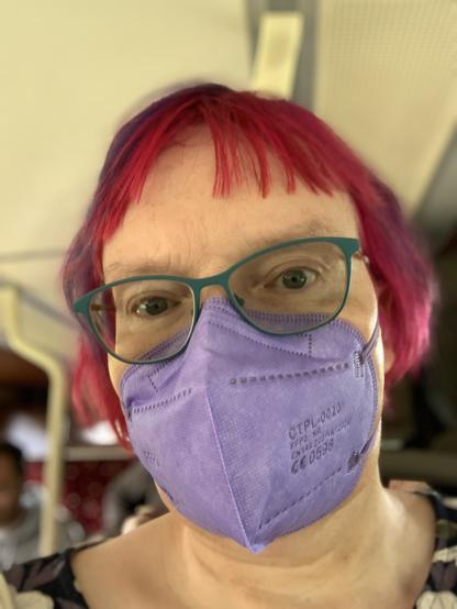 Anna is seated near the back of a bus. She is a white woman with short bobbed red hair streaked with blue. She is wearing green glasses, a lilac FFP2 face mask, and a floral print dress in non-binary Pride colours.