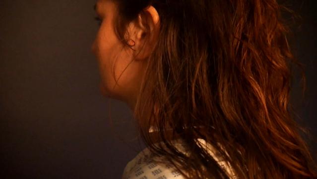 Videostill aus der Installation Mensch | Maschinen | Raum des Teenage Angst Ensembles: Kopf und Schultern einer weiblichen Person von schräg hinten. Sie hat lange, gelockte Haare, trägt ein Krankenhaushemd und steht in einem undefinierten, nebligen Raum.