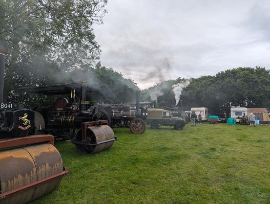 Steam engines and vintage vehicles displayed in an outdoor setting with smoke rising, trees in the background, and people walking around.