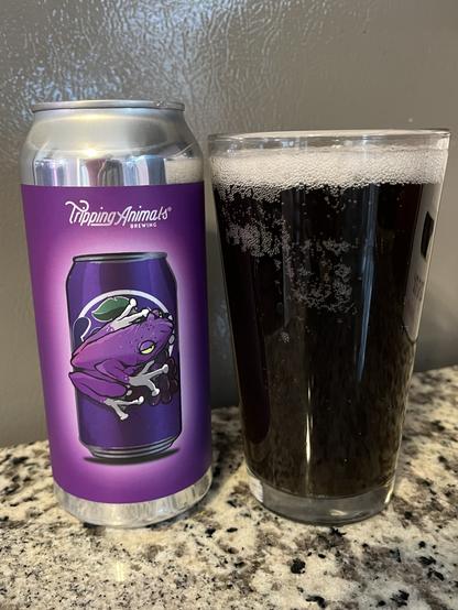 A can of beer from Tripping Animals Brewing with a purple label featuring a frog, placed next to a glass of dark beer on a granite countertop.