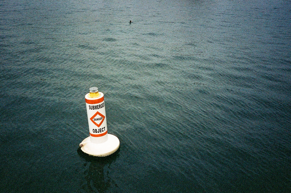 A white warning buoy with red markings and black text reading “SUBMERGED OBJECT DANGER”. There is a duck in the distance.