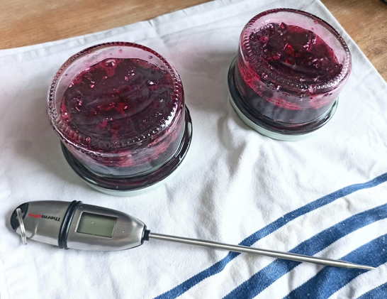 Auf einem Geschirrtuch 2 auf dem Kopf stehende Marmeladegläser mit Kirschmarmelade. Davor liegt das Thermometer