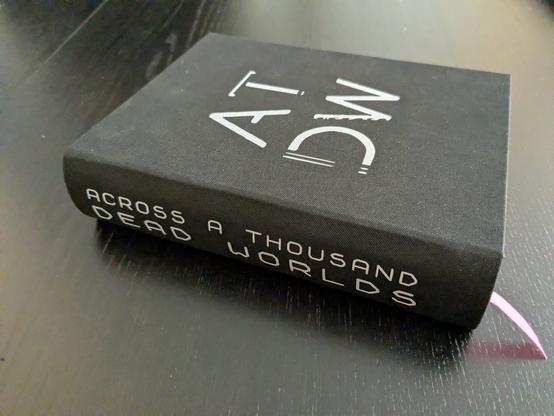 The finished book lying on a dark table. It's covered in black cloth. On the front are the letters "ADTW" in a two by two letter square, on the spine it says "Across a Thousand Dead Worlds" in two lines.