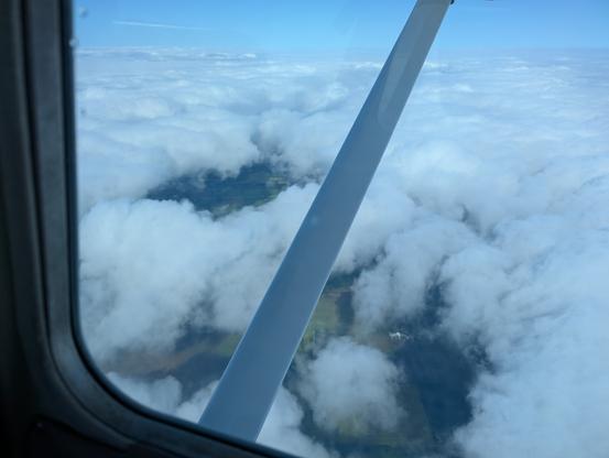 Looking out right window of a Cessna Skyhawk. The wing strut is running across the middle of the frame. We are peering down between a small gap in the clouds revealing a dreary day below.