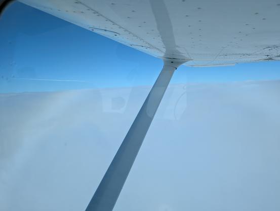 Looking out the right side of a Cessna Skyhawk. The high wing is visible taking up the top of the frame. There is a sea of clouds below all the way to the horizon and clear skies above