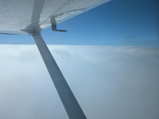 Looking out the left side of a Cessna Skyhawk, the wing and strut are visible in the frame. The plane is just above the clouds at 6000 feet. Skies above are blue and clear. On the wing the pitot tube is visible, which measures the airspeed for the pilot. It is a metal piece coming down, then forward at a 90 degree angle.