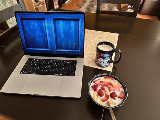 A laptop displaying a blue background with two rectangle shapes, a mug of milk on a coaster, and a bowl of cereal with strawberries on a dark table.