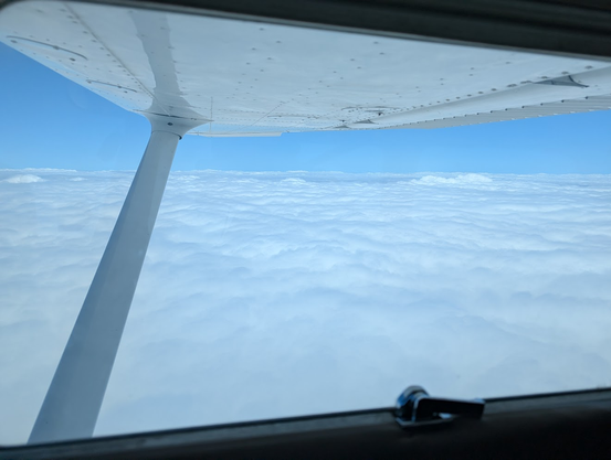 Looking out the right side of a Cessna Skyhawk. The wing is on the top of the frame, the sky is clear, and below is a solid floor of clouds.