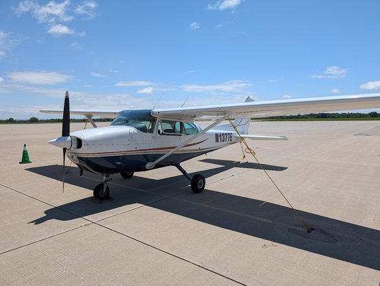 A C172 Cessna Skyhawk parked on the ground. Yellow tie down ropes secure it to its position on the concrete. Above the sky has a few clouds but it is mostly clearing