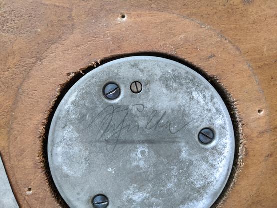 Nahaufnahme eines runden Metallteils auf das etwas mit Bleistift(?) geschrieben wurde. Es ist in einen hölzernes Gestell eingepasst.