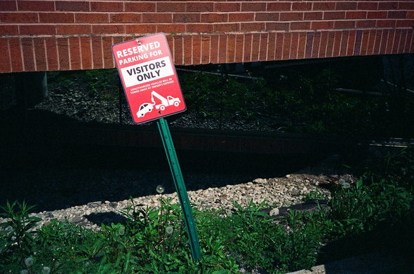 A sign at a medical building reads: reserved parking for visitors only, unauthorized vehicles will be towed away at owner’s expense. The sign is red with white printing and an illustration of a car being towed away. It is on a bent green stanchion.