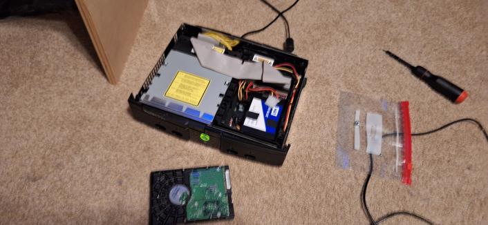 An original Xbox powered on with it's case open on the top. A hard drive is infront of the console. In the hard drive bay, there's a solid state drive electrical tapped in place.