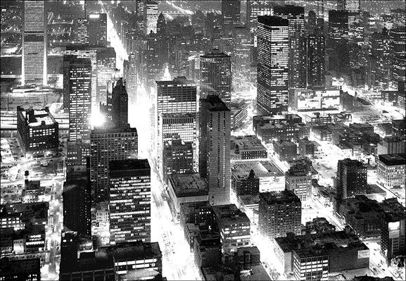 Monochrome view of skyscrapers and city streets from above at night, illuminated by long exposure so streets and especially one main street are nearly all white.