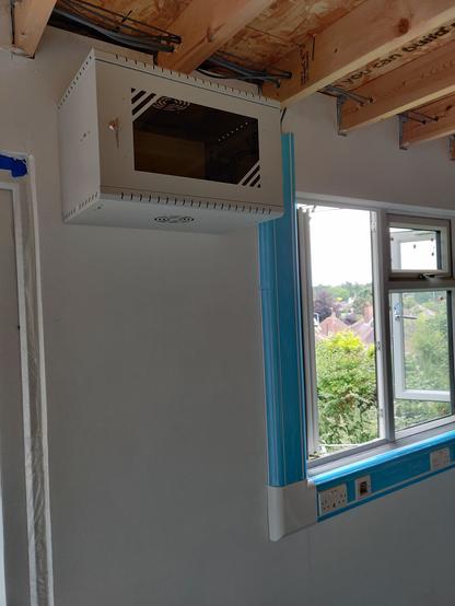 a white office wall with some open windows and trunking running below and up one side of the window. At the top of the wall is a small networking cabinet that looks like a microwave.