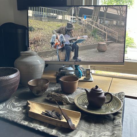 A small tea ceremony laid out with handmade teaware on fabric patterned with green Chinese dragons. A wooden teascoop holds a few grams of an aged puerh tea, almost 25 years in age. 

In the background on the monitor, the Japanese guitarist Taku Sugimoto plays an archtop guitar. He performs seated, in an urban setting in the open air, two microphones positioned in front of the guitar’s soundhole.