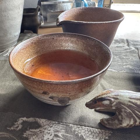 A small teabowl with tea in it. The bowl has an abstract stylised leaf painting on the front. There is a small jug in the background and the front half of a ceramic badger to the right of the frame.