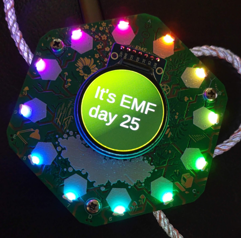 Photo of an EMF Tildagon badge showing the output of the EMF_Day badge app. The text reads "It's EMF day 25" on the Tildagon's round central screen, and the LEDs around the edge of the badge are illuminated in a rainbow pattern.