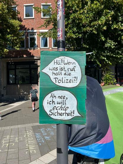 Ein Pappschild mit zwei Sprechblasen auf grünem Hintergrund. 
Erste Sprechblase: "Hä? Wenn was ist, ruf halt die Polizei?!"
Zweite Sprechblase: "Äh nee, ich will echte Sicherheit!"