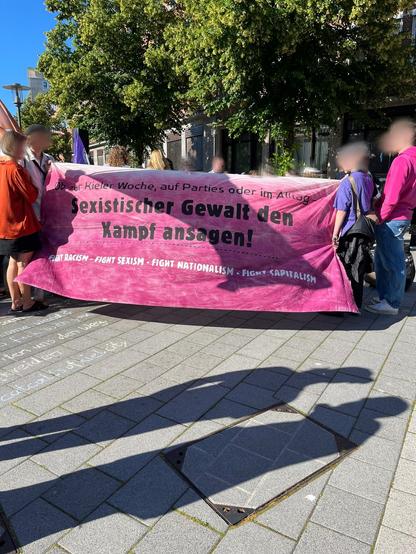 Mehrere Menschen halten ein Transparent mit einem Farbverlauf weiß zu pink darauf, davor die Schrift: "Ob zur Kieler Woche, auf Parties oder im Alltag: Sexistischer Gewalt den Kampf ansagen! Fight Racism - Fight Sexism - Fight Nationalism - Fight Capitalism".