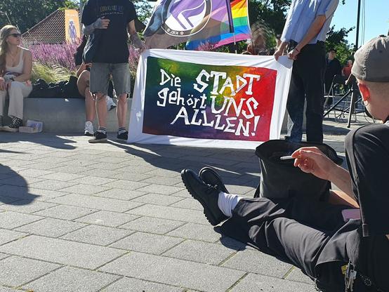 Vor einer Regenbogenflagge und der Flagge der Feministischen Antifa Kiel ein kleines Transparent, darauf in weiß vor einem regenbogenfarbenen Hintergrund: "Die Stadt gehört uns allen!"