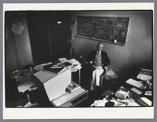 The image shows an individual seated in a cluttered office environment. The person appears to be wearing glasses and is dressed in casual attire with a tie, suggesting a formal or professional setting. There are various items scattered around the room, including papers, books, and other objects that contribute to a sense of disarray. The room is dimly lit, which could indicate an older photograph or simply a preference for low light conditions by the individual. The background features a desk with more clutter and shelves holding additional materials. There are no visible texts within the image. The overall atmosphere suggests a scene from daily life, possibly in an office where the person is engaged in some work or study.