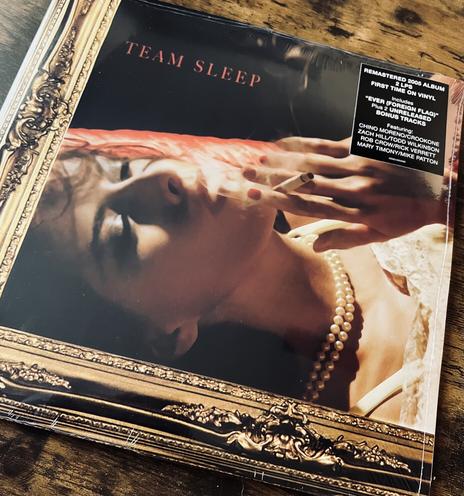 A vinyl copy of The self titled album by the band Team sleep. A woman smokes a cigarette. She is laying down. It has a gold frame around it