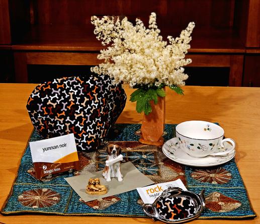 "A tea tableau in somber, autumnal tones depicting dogs awaiting their person's return. One looks hopeful, holding a bone in its mouth, the other two seem anxious, clinging together. There is a cloud of ivory ocean spray flowers, rather like a funeral arrangement, & a packet of Rock Sugar signifying the bedrock steadiness & distilled, crystalline purity of canine devotion. The tea is Yunnan Noir, for the long night of the soul that is separation."
