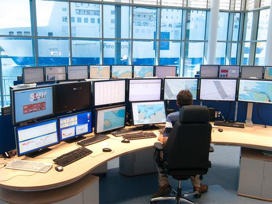 Ein Nautiker hat in der Verkehrszentrale auf Monitoren den Schiffsverkehr im Blick. Vor dem Fester passiert ein großes Schiff die Verkehrszentrale.