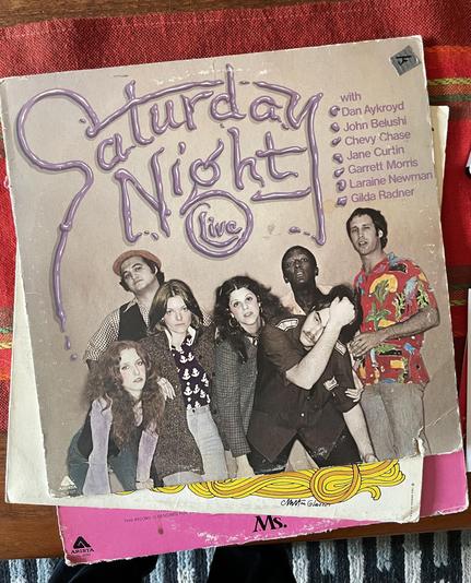 Photo of three record sleeves on a table. On top is a cover for “Saturday Night Live” showing members of the 1976 cast and purple lettering that looks like it is liquid extracted from a tube.