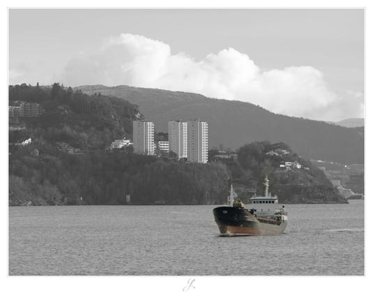 Landscape shot from the deck of a ship looking towards the shoreline. A ship is sailing towards us between us and the shoreline. In the background we can see the steep bank with high-rise buildings and surrounding forest. In the background we see a gentle, wooded range of hills. Above it are large clouds in the sky. The picture is almost black and white and has just a soft color tone for the ship.

AI disclaimer: Using my work, its meta data, written or derived description to create media with or train AI based systems is prohibited.