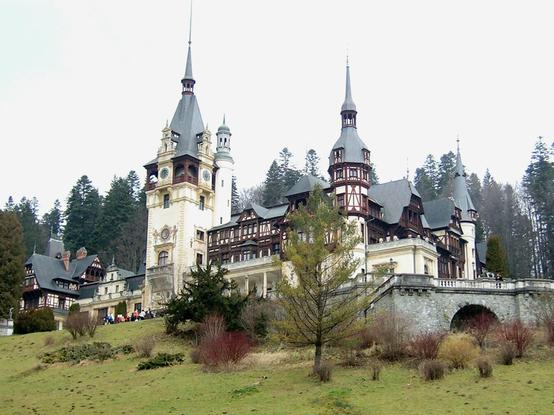 A large, ornate building with multiple spires and intricate architectural details. The structure combines various styles, including a central tower with a conical roof, several other towers featuring pointed spires, and balconies. The surrounding area includes grassy slopes with leafless trees.