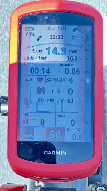 Urządzenie Garmin wyświetlające różne wskaźniki rowerowe, w tym prędkość (14,3 km/h), temperaturę (38,7°C), czas (11:32), RPM (0), tętno (89 uderzeń na minutę) i odległość (0,06 km). W lewym, dolnym narożniku informacja o zagrożeniu na drodze.