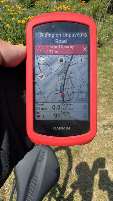 A handheld Garmin GPS device showing a map with a "Hazard Nearby" warning. A bicycle saddle and a garden with yellow flowers are in the background.

Ręczne urządzenie Garmin GPS wyświetlające mapę z ostrzeżeniem "Zagaranie w pobliżu". W tle znajduje się siodełko rowerowe i ogród z żółtymi kwiatami.