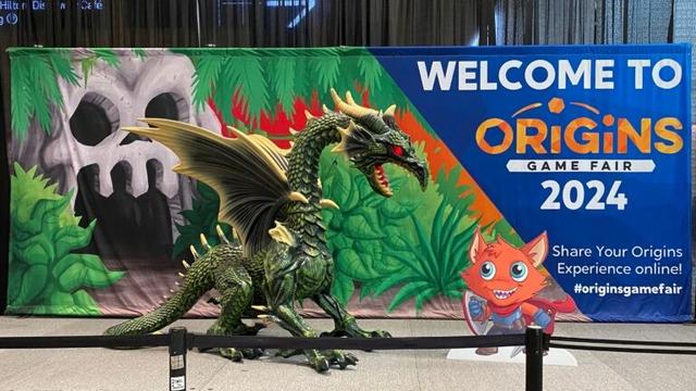 Animatronic dragon and a standee of the Origins Game Fair mascot, "Crit Cat", in front of a banner reading "Welcome to Origins Game Fair 2024". The banner shows a cave entrance carved to look like a skull.