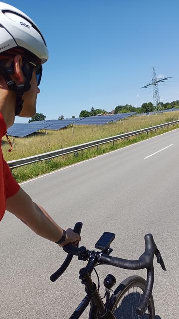 Im Vordergrund fährt ein Rennradfahrer mit weißem Helm, orangenen Trikot und schwarzem Rennrad auf einer Landstraße. Im Hintergrund ein großes Solarfeld und ein Strommast. Der Himmel ist blau und wolkenlos.