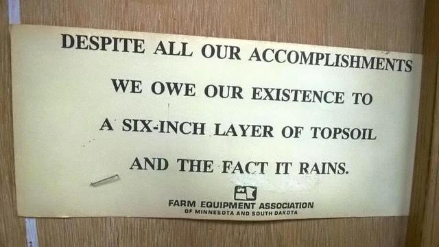 Photo of a sign that reads: "Despite all our accomplishments we owe our existence to a six-inch layer of topsoil and the fact that it rains." The sign is printed on aged cardstock with "Farm Equipment Association of Minnesota and South Dakota" at the bottom, stapled to a wooden door.